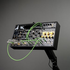 Könnte beinhalten: Schwarzer modularer Synthesizer mit zahlreichen Knöpfen, Tasten und Anschlüssen, verbunden durch grüne und weiße Kabel. Der Synthesizer ist auf einem schwarzen Ständer vor grauem Hintergrund montiert. Goldfarbene Knöpfe und verschiedene Textbeschriftungen sind sichtbar.
