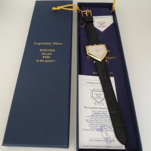May include: A home plate-shaped wristwatch with a gold-tone case and a black leather strap, presented in a navy blue box. The box lid features the text Legendary Man WATCHES that put YOU in the game. Includes a certificate of authenticity.
