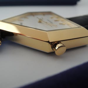 May include: A gold-tone watch with a pentagonal case and a white dial featuring gold-tone hour markers and hands. The watch includes a black leather strap attached with gold-tone screws and a side-mounted crown for time adjustment.
