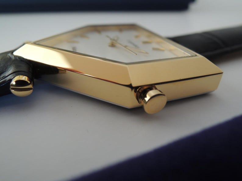 May include: A gold-tone watch with a pentagonal case and a white dial featuring gold-tone hour markers and hands. The watch includes a black leather strap attached with gold-tone screws and a side-mounted crown for time adjustment.