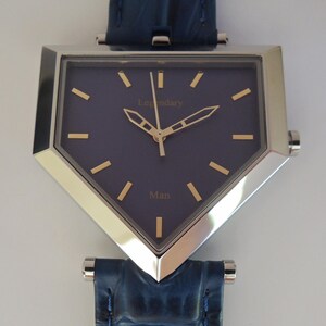 May include: A shield-shaped wristwatch with a dark blue dial, gold-tone hands, and gold-tone hour markers. The dial features the text Legendary and Man in gold lettering. The watch has a polished silver-tone metal case and a dark blue textured leather strap.