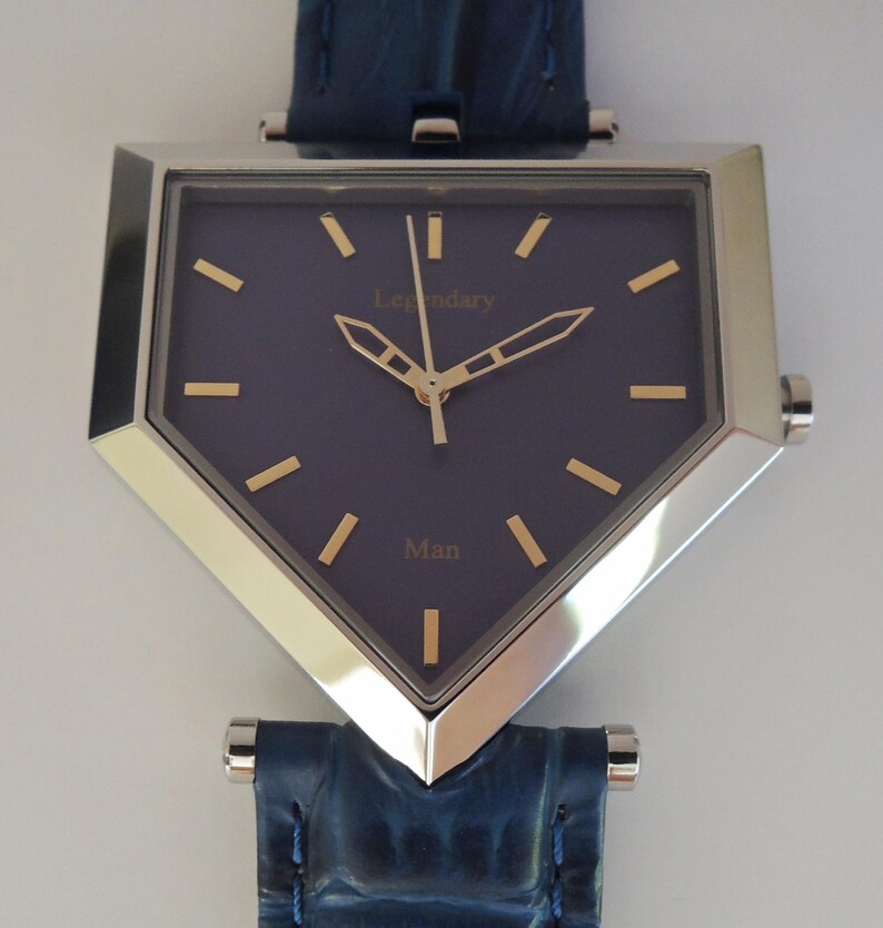 May include: A shield-shaped wristwatch with a dark blue dial, gold-tone hands, and gold-tone hour markers. The dial features the text Legendary and Man in gold lettering. The watch has a polished silver-tone metal case and a dark blue textured leather strap.
