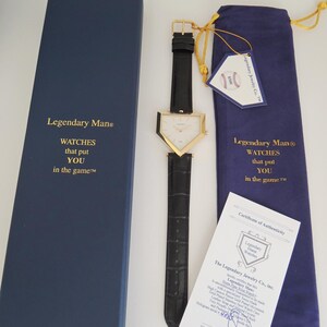 May include: A gold-tone triangular watch with a black leather strap, presented with a blue box, a blue drawstring pouch, a baseball-themed tag, and a certificate of authenticity. The box and pouch feature the text Legendary Man WATCHES that put YOU in the game.