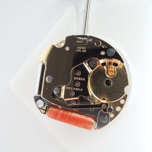 May include: A gold-toned Ronda watch movement featuring the text SWISS EOL R9, RONDA, and FIVE 5 JEWELS. The mechanism includes a prominent orange copper coil, various metallic gears, and small screws on a circular plate with a metallic stem.