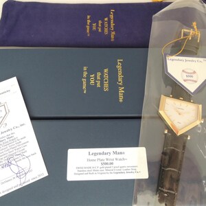 May include: A gold-toned, home plate-shaped wristwatch with a dark brown leather strap, presented in a blue box with a matching pouch, a certificate of authenticity, and a tag featuring a baseball graphic and the text Legendary Jewelry Co.