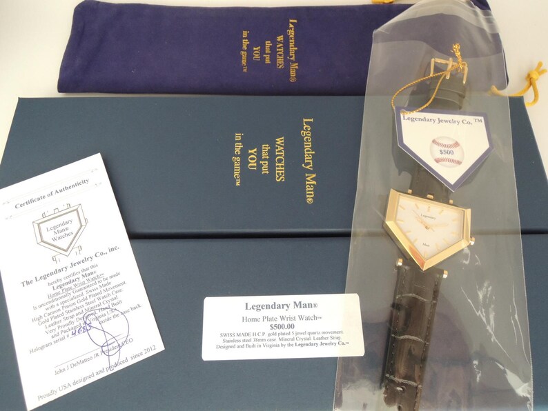 May include: A gold-toned, home plate-shaped wristwatch with a dark brown leather strap, presented in a blue box with a matching pouch, a certificate of authenticity, and a tag featuring a baseball graphic and the text Legendary Jewelry Co.