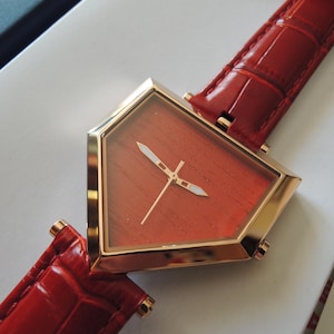 May include: A pentagonal wristwatch with a gold-tone metal case and a red wood-grain dial. The watch features thin gold-tone hands with white accents and is attached to a glossy red leather strap with a textured, crocodile-embossed pattern.
