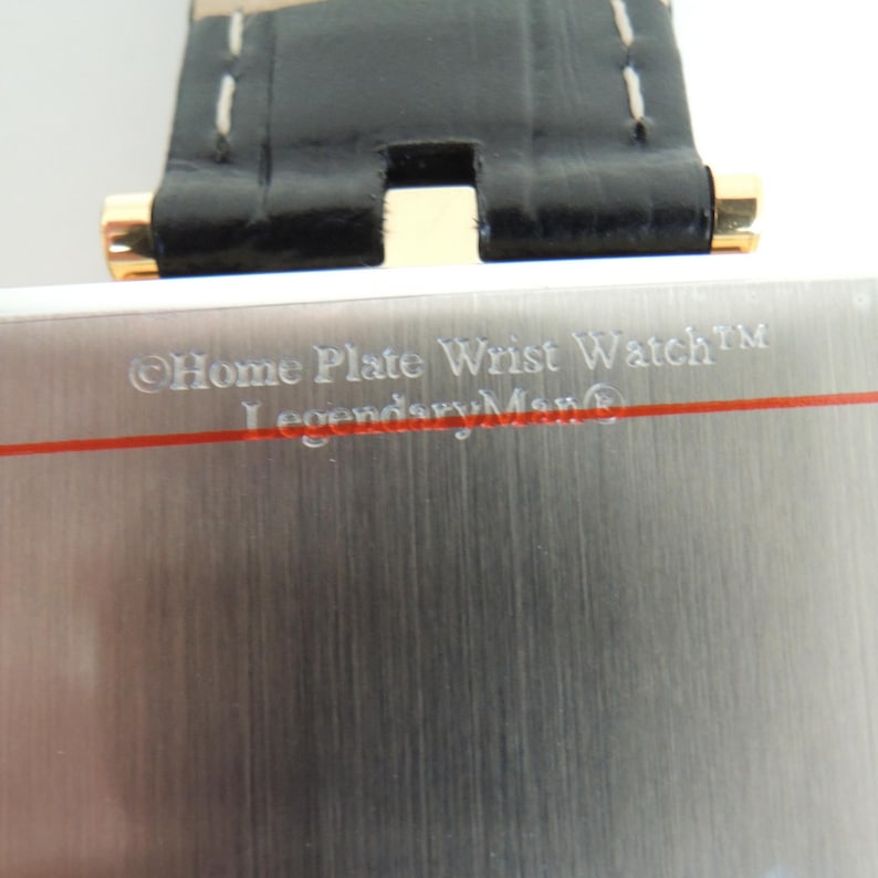 May include: A wristwatch with a brushed silver-tone metal case featuring a horizontal red line and the engraved text Home Plate Wrist Watch and LegendaryMan. The watch includes a black leather strap with white stitching and gold-tone hardware.