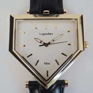 May include: A triangular-shaped wristwatch with a gold-tone metal case and a white dial. The dial features the text 'Legendary' and 'Man' in black, with gold-tone hour markers and hands. It is attached to a black textured leather strap.