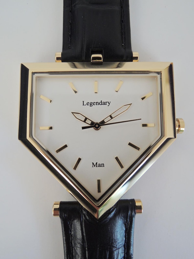 May include: A triangular-shaped wristwatch with a gold-tone metal case and a white dial. The dial features the text 'Legendary' and 'Man' in black, with gold-tone hour markers and hands. It is attached to a black textured leather strap.