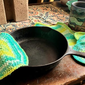 May include: A black cast iron skillet with a handle, alongside several crocheted pot holders and scrubbers in shades of green, yellow, and white. A mug with a landscape design is visible in the background.