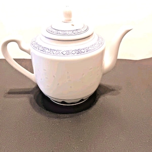May include: White porcelain teapot with a lid and a curved handle. The teapot features a blue geometric pattern around the lid and base. The teapot is sitting on a dark surface.