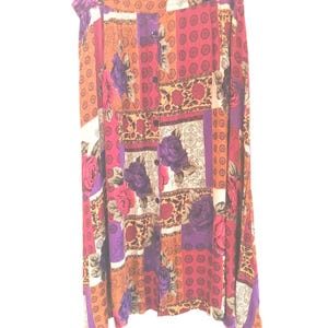 May include: A long, button-down skirt with a patchwork design. The skirt features a mix of floral and geometric patterns in shades of purple, pink, orange, and cream. The skirt is hanging on a white hanger.