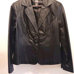 May include: A black leather blazer featuring a notched lapel, long sleeves, and a three-button front closure. The jacket includes a horizontal seam detail across the waist and a smooth interior lining, displayed on a wooden hanger.