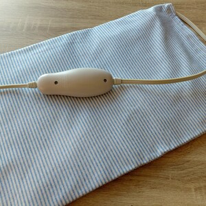 May include: A cream-colored heating pad with a white control unit and cord, resting on a blue and white striped fabric. The pad is designed for therapeutic use and is likely used for pain relief or relaxation.