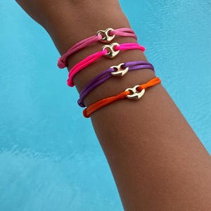May include: A collection of colorful bracelets in shades of pink, purple, and orange. Each bracelet features a gold-colored clasp. The bracelets are displayed on a person's wrist, with a blue water background.