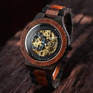 May include: A wooden wristwatch with a black face and visible gold-colored gears. The watch has a brown wooden bezel and a two-tone wooden band. The watch is displayed on a wooden surface.