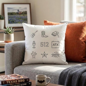 May include: A white square throw pillow with black line art of a guitar, cowboy boot, baseball glove, microphone, the number 512, a sign that says ATX, a cactus, a star, and a bridge. The pillow is on a gray couch with orange and rust-colored pillows.