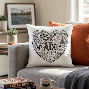 May include: White throw pillow with a black heart-shaped design featuring iconic Austin, Texas symbols, including a guitar, bats, and the Texas State Capitol. The pillow is on a gray couch with an orange pillow.