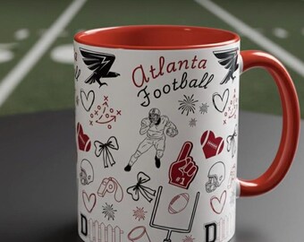 Puede incluir: Una taza de cerámica blanca con interior y asa rojos, con ilustraciones en negro y rojo relacionadas con el fútbol. La taza tiene las palabras "Atlanta Football" en una fuente de escritura.