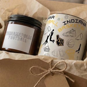 Könnte beinhalten: Eine Geschenkbox mit einer Kerze und einer Tasse. Die Kerze hat einen schwarzen Deckel und ein Etikett mit der Aufschrift "Indianapolis Football". Die Tasse ist weiß mit schwarzen Akzenten und Basketball-Grafiken.