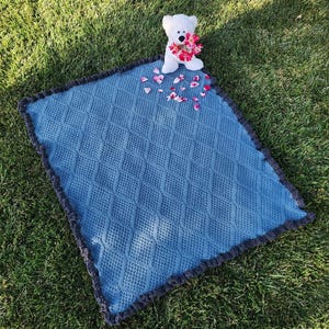 Puede incluir: Una manta de bebé tejida de color azul con un patrón de diamantes y un borde gris oscuro. Un osito de peluche blanco con una corona floral está sentado sobre la manta, rodeado de pétalos de flores rosas esparcidos. La manta está sobre hierba verde.