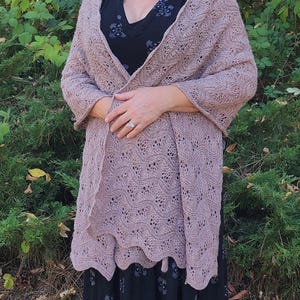 May include: A mauve-colored, open-front shawl with a delicate, openwork design. The shawl is draped over a black dress with a floral pattern. The shawl's edges have a scalloped trim, adding a touch of elegance to the knitted garment.