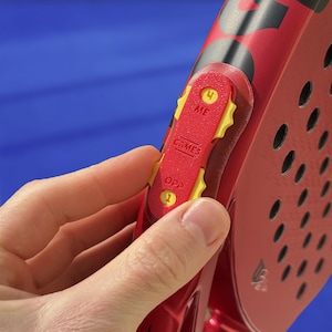 Puede incluir: Primer plano de una raqueta deportiva roja y negra con superficie perforada. Se ve un mecanismo de ajuste rojo y amarillo, con los textos "ME" y "OPP" y los números 1 y 4. La raqueta se sostiene sobre un fondo azul.