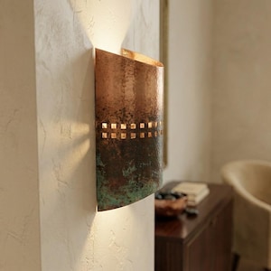 May include: A copper wall sconce with a half-cylinder shape. The sconce has a textured, aged appearance with a mix of brown, green, and copper tones. A row of square cutouts allows light to shine through.