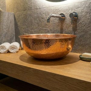 May include: A hammered copper sink bowl sits on a wooden countertop in a bathroom setting. The sink is round with a textured surface and a shiny copper finish. Rolled white towels and soap dishes are also visible.