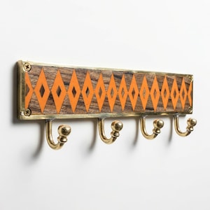 May include: A wooden wall-mounted coat rack with five brass hooks. The wooden board features a geometric pattern of orange diamonds on a brown background, framed by brass edging. The hooks are curved and have a ball-shaped end.