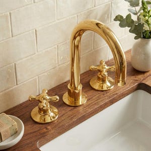 May include: A polished gold bathroom faucet set featuring a high-arched spout and two separate cross-handle controls. The fixture is mounted on a dark wood countertop next to a white ceramic sink basin and a small bar of soap in a dish.