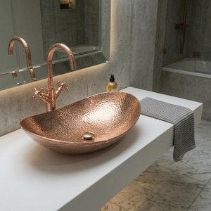 May include: A copper-coloured, hammered metal oval sink sits on a white countertop. A matching copper tap with cross handles is mounted behind the sink. A grey towel hangs on the counter. The bathroom has a marble tile floor.