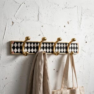 Puede incluir: Un perchero decorativo de pared con cuatro ganchos de latón. El perchero presenta un patrón de diamantes en blanco y negro, ribetes dorados y tornillos rojos. Una bufanda beige y un bolso de mano cuelgan de los ganchos.