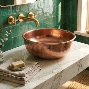 Puede incluir: Un lavabo de cobre martillado sobre una encimera de mármol, con un grifo de latón y azulejos verdes. Toallas beige dobladas y una pastilla de jabón completan la escena del baño. El lavabo es el elemento principal.
