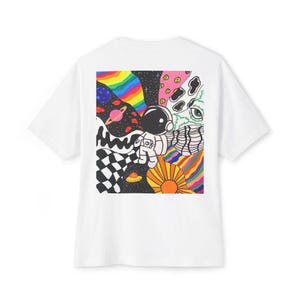 Puede incluir: Camiseta blanca con un diseño psicodélico y colorido en la espalda. El diseño incluye un astronauta, planetas, arcoíris y un patrón de tablero de ajedrez. La obra de arte tiene un estilo retro y de dibujos animados.