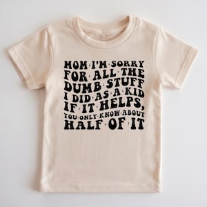 May include: A beige short-sleeved t-shirt with the text "MOM I'M SORRY FOR ALL THE DUMB STUFF I DID AS A KID IF IT HELPS, YOU ONLY KNOW ABOUT HALF OF IT" in black. The shirt is made of a soft material.