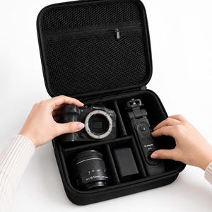 May include: A black, square, zipped camera carrying case with an open lid featuring a mesh storage pocket. The interior contains a black Canon EOS R7 camera body, an EFS 18-55mm lens, a rectangular battery pack, and a black Canon BR-E1 remote control.