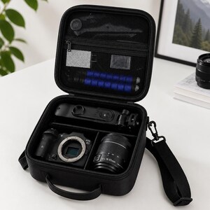Op de afbeelding: Een zwarte, vierkante cameratas met ritssluiting en een open deksel. De binnenkant is voorzien van een gaasvak met kleine accessoires en een hoofdvak met verdelers voor een camerabody, een lens en een zwarte statiefbevestiging.