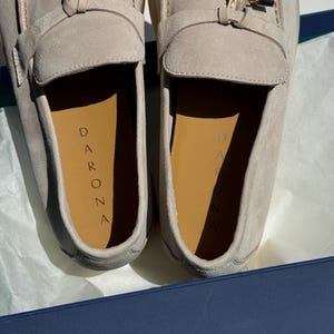 May include: A pair of light beige suede loafers featuring a gold-toned metal hardware accent on the vamp. The interior insoles are tan and clearly display the brand name DARONA printed in black lettering. The shoes are presented inside a box.