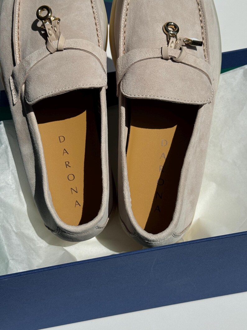 May include: A pair of light beige suede loafers featuring a gold-toned metal hardware accent on the vamp. The interior insoles are tan and clearly display the brand name DARONA printed in black lettering. The shoes are presented inside a box.
