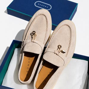 May include: A pair of beige suede loafers featuring a gold-toned metal charm on the vamp and a tan insole with the brand name DARONA printed in black. The shoes are presented in a navy blue box with the brand name DARONA on the lid.