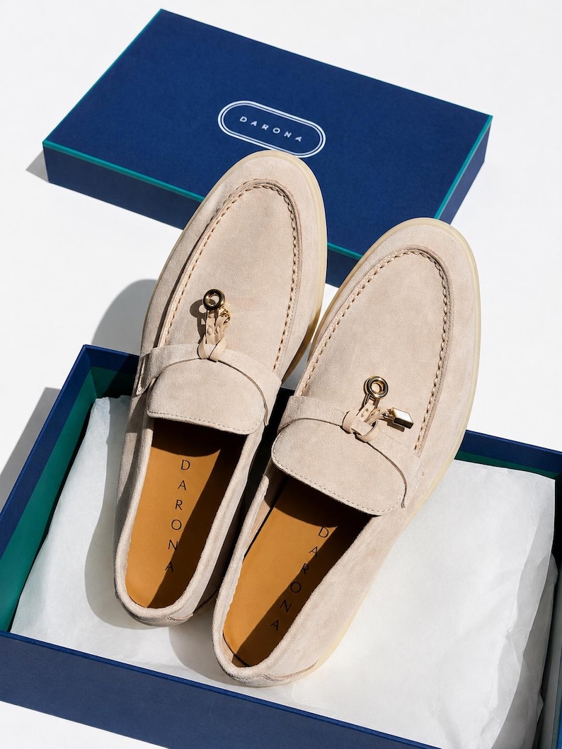 May include: A pair of beige suede loafers featuring a gold-toned metal charm on the vamp and a tan insole with the brand name DARONA printed in black. The shoes are presented in a navy blue box with the brand name DARONA on the lid.