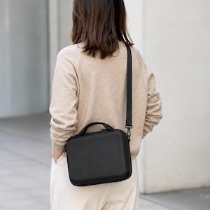 K&ouml;nnte beinhalten: Eine schwarze, rechteckige Umh&auml;ngetasche mit einem oberen Griff und einem abnehmbaren Riemen. Die Tasche wird quer &uuml;ber dem K&ouml;rper getragen. Die Person tr&auml;gt einen beigen Pullover und eine wei&szlig;e Hose.