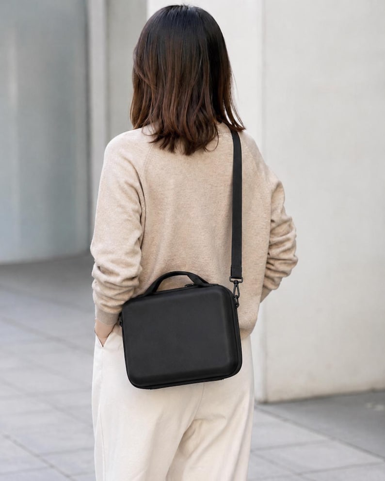 K&ouml;nnte beinhalten: Eine schwarze, rechteckige Umh&auml;ngetasche mit einem oberen Griff und einem abnehmbaren Riemen. Die Tasche wird quer &uuml;ber dem K&ouml;rper getragen. Die Person tr&auml;gt einen beigen Pullover und eine wei&szlig;e Hose.
