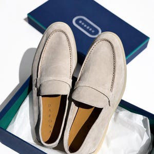 May include: A pair of light beige suede loafers with a classic penny strap design. The interior of the shoes features a tan lining with the brand name DARONA printed in black. The loafers are presented inside a dark blue box with white tissue paper.