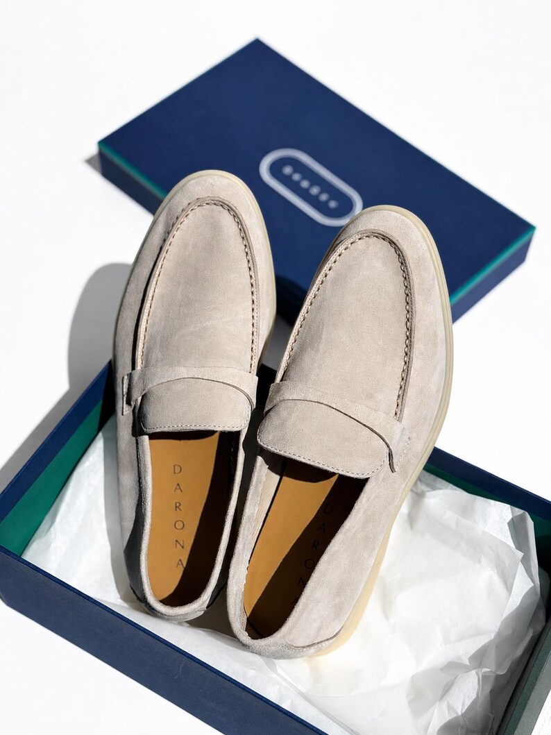 May include: A pair of light beige suede loafers with a classic penny strap design. The interior of the shoes features a tan lining with the brand name DARONA printed in black. The loafers are presented inside a dark blue box with white tissue paper.