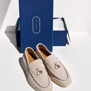 May include: A pair of beige suede loafers with a gold-toned metal charm featuring a square and circular design on the vamp. The shoes are positioned in front of a rectangular blue box with the brand name DARONA printed in white text on the lid.