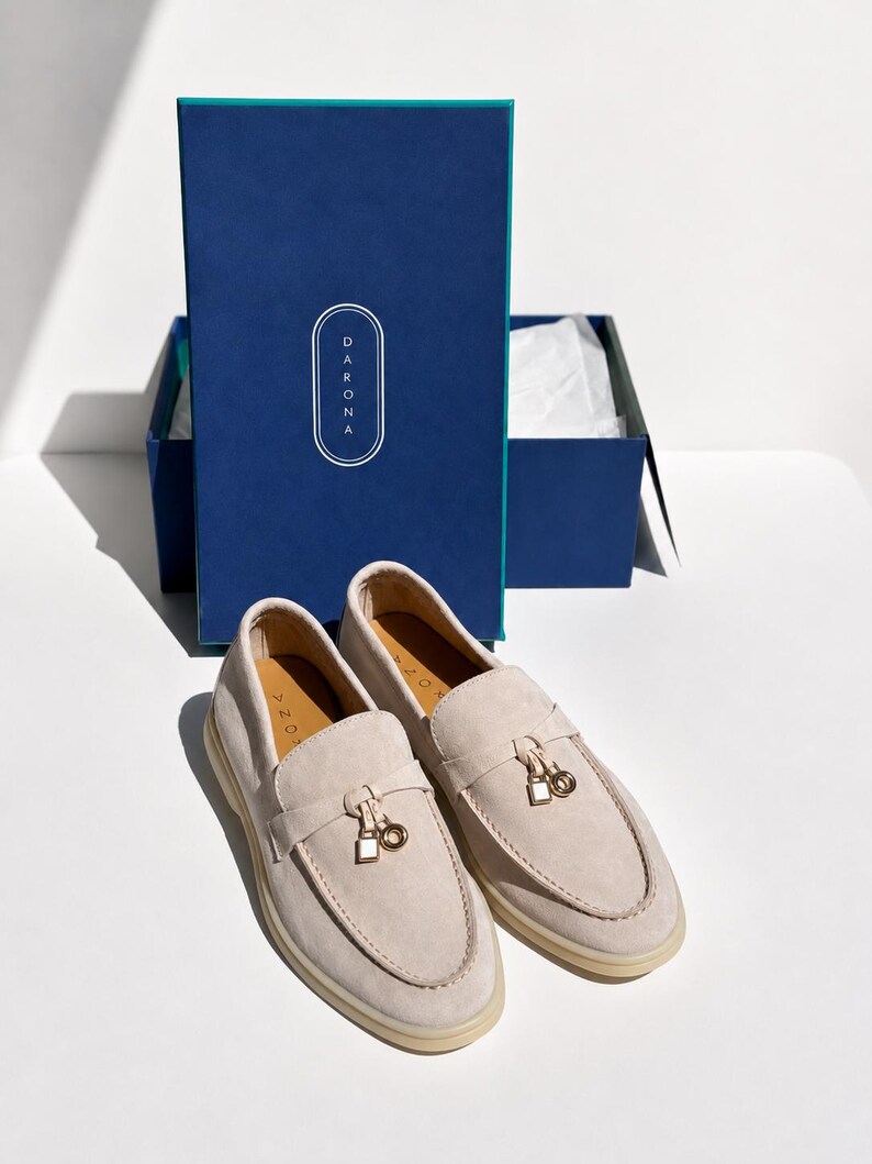 May include: A pair of beige suede loafers with a gold-toned metal charm featuring a square and circular design on the vamp. The shoes are positioned in front of a rectangular blue box with the brand name DARONA printed in white text on the lid.