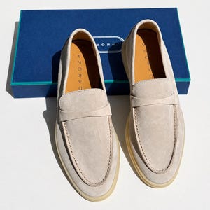 May include: A pair of beige suede loafers with a classic penny strap across the vamp, featuring light-colored rubber soles and tan leather insoles stamped with the brand name DARONA, positioned in front of a blue rectangular shoe box.
